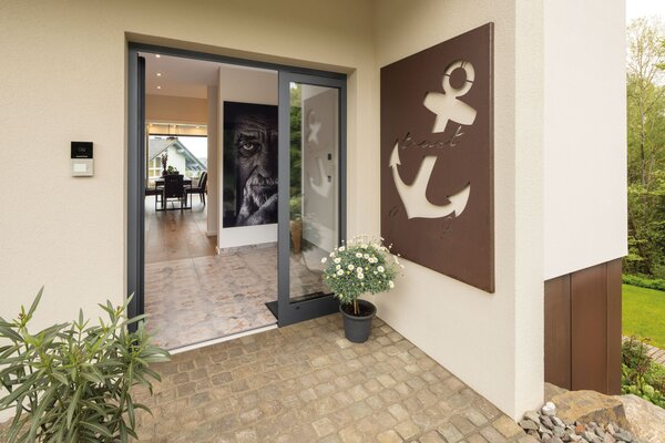 Moderner Hauseingang eines FingerHaus-Hauses mit dunkler Tür, bepflanztem Eingangsbereich und stilvoller Wanddekoration in Ankerform aus Metall.