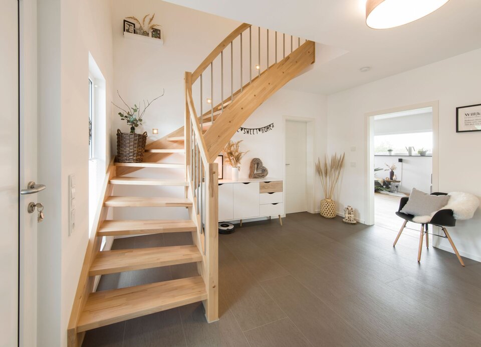 Heller Flur mit offener Holztreppe und geschwungenem Geländer. Dekoration mit Pflanzen, Körben und Sesseln, brauner Bodenbelag und weiße Wände.