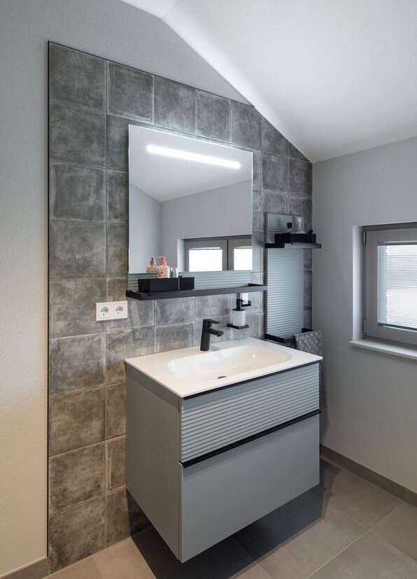 Modernes Badezimmer im FingerHaus ARTIS 400 Musterhaus Schwarzwald mit Waschtisch, großem Spiegel mit Beleuchtung und grauen Fliesen.