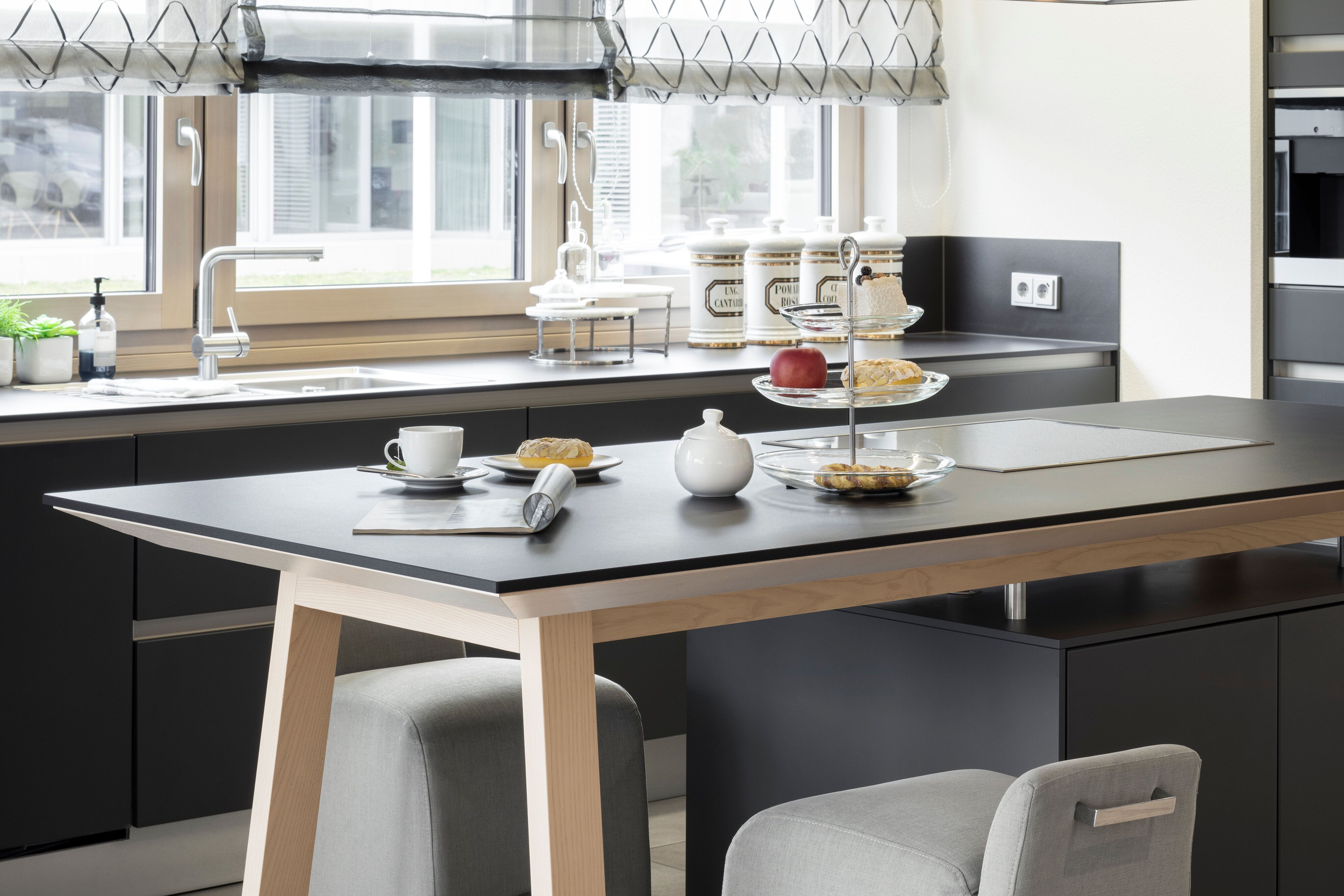 Moderne Küche im Musterhaus NEO 312 in Bad Vilbel mit dunkler Arbeitsfläche, heller Holztischinsel und zwei gepolsterten Hockern. Auf der Theke stehen Geschirr, Obst und Gebäck, während durch große Fenster viel Tageslicht einfällt.