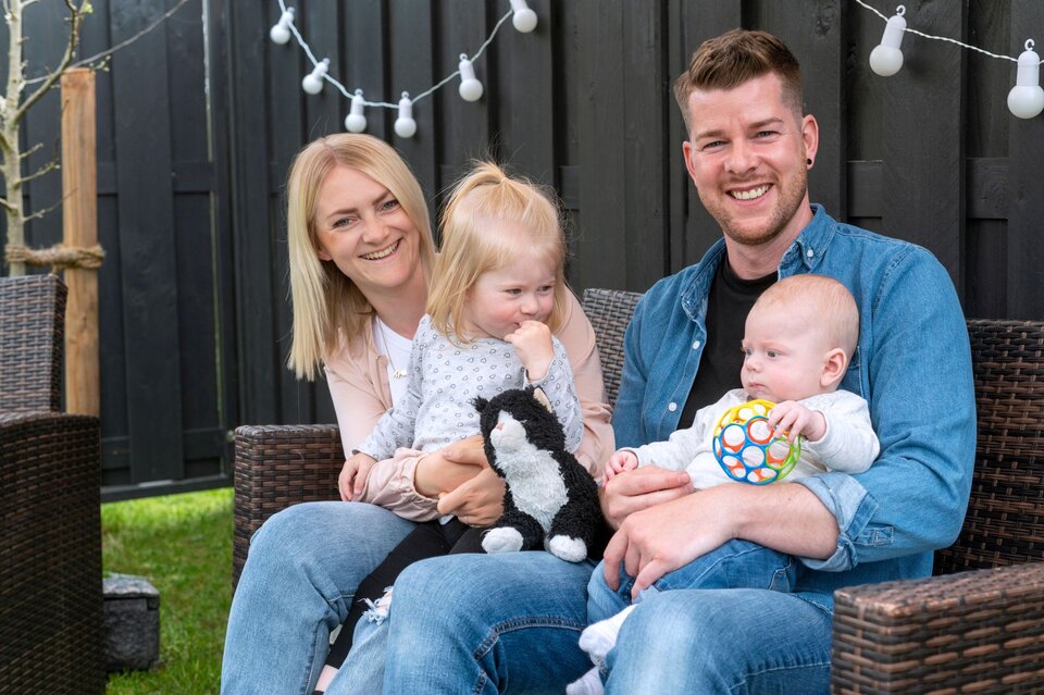Ein Paar sitzt mit zwei kleinen Kindern auf Gartenmöbeln. Das ältere Kind hält ein Stofftier, das Baby eine bunte Spielkugel. Im Hintergrund ist ein Garten mit Rasen, Holzzaun und Lichterkette zu sehen.