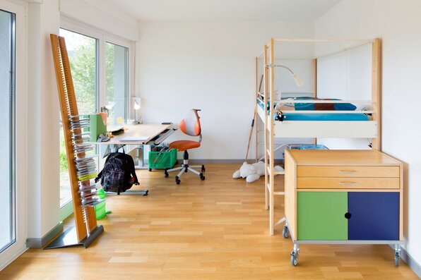 Helles Kinderzimmer mit Hochbett, farbenfrohem Möbelstück und Schreibtisch am großen Fenster mit Blick ins Grüne.