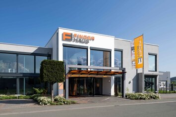 Außenansicht der FingerHaus-Zentrale in Frankenberg (Eder) mit moderner, zweigeschossiger Architektur, großflächigen Glasfassaden, weiß-grauer Fassade, überdachtem Eingangsbereich, FingerHaus-Logo an der Fassade und Fahne vor dem Gebäude bei Tageslicht.