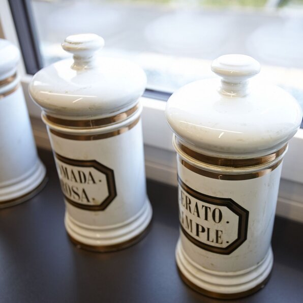 Nahaufnahme von dekorativen Keramikgefäßen mit Deckel und goldenen Akzenten, die auf einer Fensterbank stehen. Die Gefäße tragen schwarze Beschriftungen im Vintage-Stil.