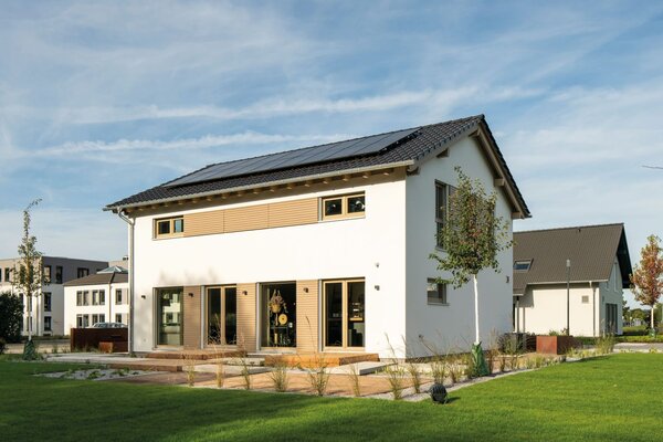 Außenansicht des FingerHaus-Musterhauses SENTO 500 in Frankenberg mit weißer Putzfassade, dunkel gedecktem Satteldach und Photovoltaikmodulen; bodentiefe Fenster zur Terrasse, große Rasenfläche, bepflanzte Beete und weitere Gebäude im Hintergrund unter blauem Himmel.