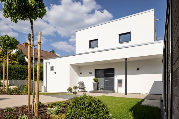 Moderner Eingangsbereich eines frei geplanten FingerHaus im Bauhaus-Stil mit klaren Linien, weißer Fassade und dunkler Haustür. Überdachung, Vorgarten mit Sträuchern und gepflasterte Wege ergänzen das Design.