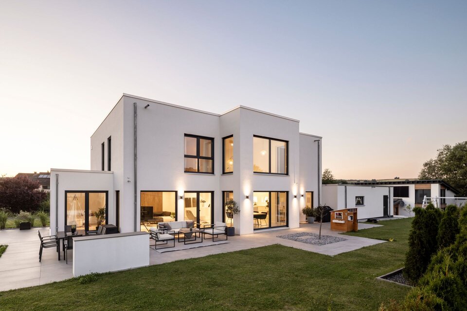 Modernes zweigeschossiges FingerHaus mit klarer, kubischer Architektur, großen Fensterflächen und stilvoll beleuchteter Terrasse in der Abenddämmerung.