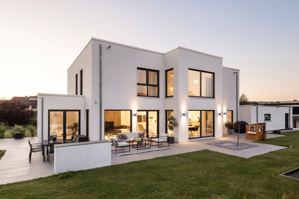 Modernes zweigeschossiges FingerHaus mit klarer, kubischer Architektur, großen Fensterflächen und stilvoll beleuchteter Terrasse in der Abenddämmerung.