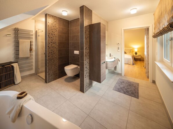 Modernes Badezimmer im Musterhaus NEO in Wuppertal mit freistehender Badewanne, ebenerdiger Dusche und stilvollen Fliesen in Holz- und Naturtönen.