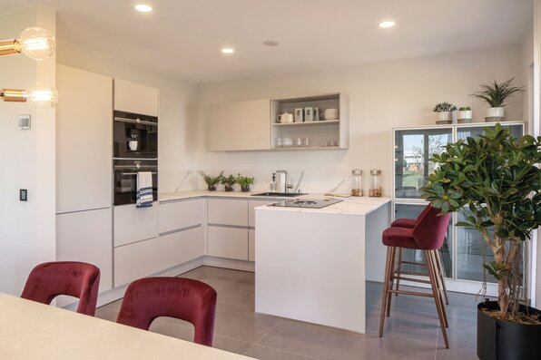 Stilvolle Küche im Musterhaus MEDLEY 300 B in Frankenberg mit moderner Kücheninsel, weißen Fronten und eleganten roten Barhockern.