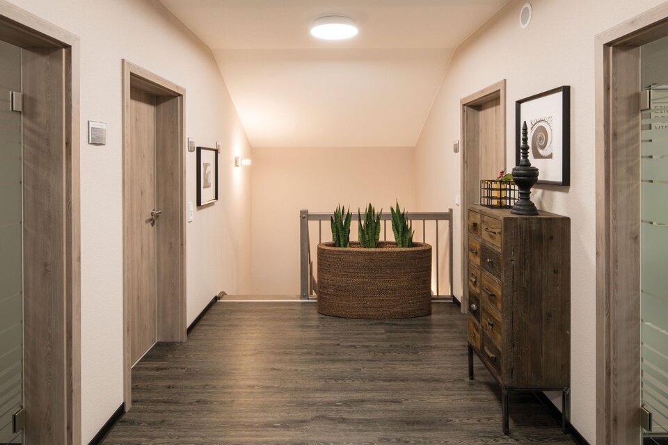 Obergeschoss-Flur mit dunklem Boden, Holztüren und Kommode mit Dekoration im Musterhaus UNO Leipzig. Am Ende des Flurs steht ein rundes Pflanzgefäß mit grünen Pflanzen, Wandbilder und Deckenleuchte ergänzen die Gestaltung.