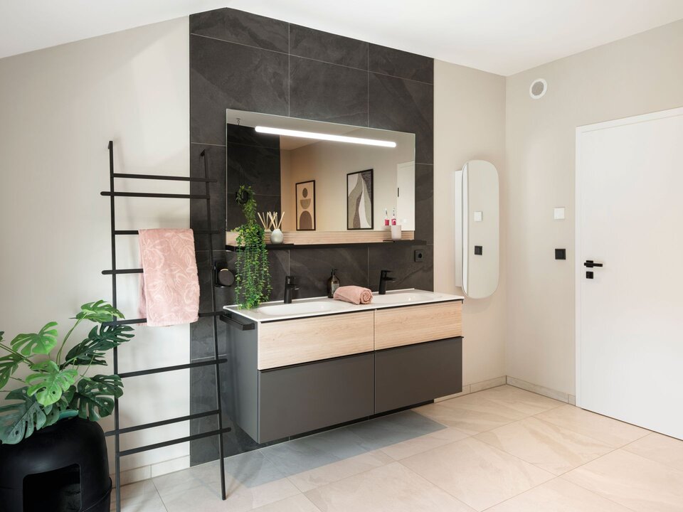 Modernes Badezimmer mit Doppelwaschtisch in Holz und Grau, großem Spiegel mit Beleuchtung und dunkler Fliesenwand. Links steht eine schwarze Handtuchleiter mit rosa Handtuch und eine grüne Zimmerpflanze.
