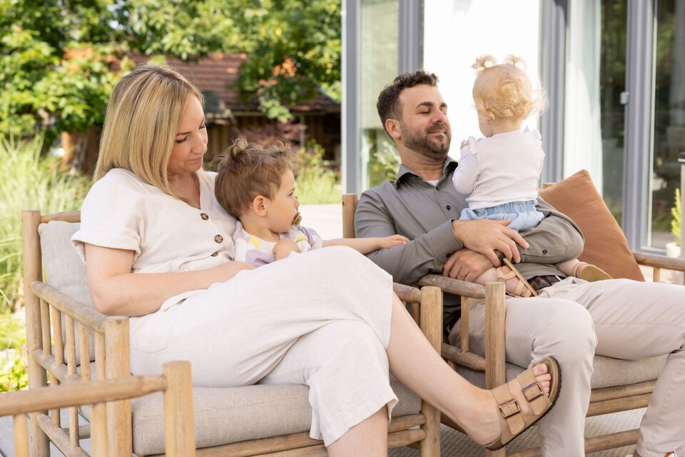 Familie entspannt auf der Terrasse eines frei geplanten FingerHaus, gemütlich sitzend auf Holzstühlen mit hellem Polster und Blick in den sonnigen Garten.
