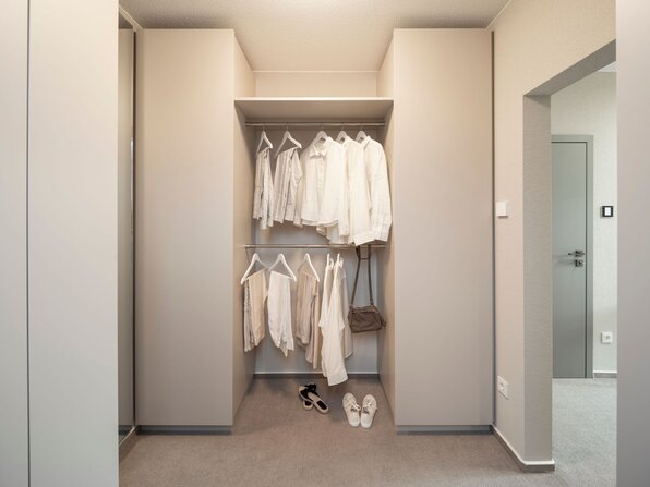 Offene Ankleide im Musterhaus JUNO 300 in Mülheim-Kärlich mit modernen Einbauschränken und hängender Kleidung in neutralen Farben.