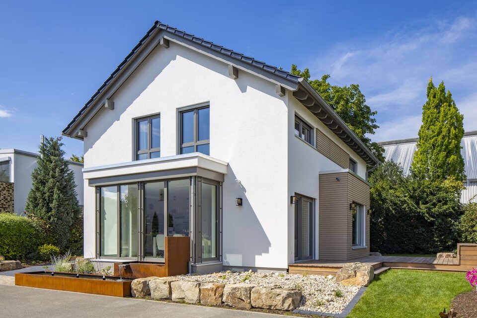 Außenansicht des FingerHaus MEDLEY Musterhauses in Mannheim mit Wintergarten, Holzakzenten und moderner Gartenanlage bei Sonnenschein.