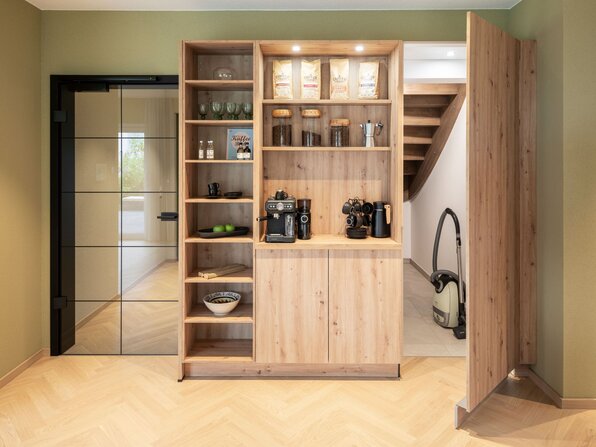 Praktischer Abstellraum im Musterhaus JUNO 300 in Mülheim-Kärlich, geschickt integriert hinter einer Holzschrankfront mit offenem Regal und Kaffeemaschine.
