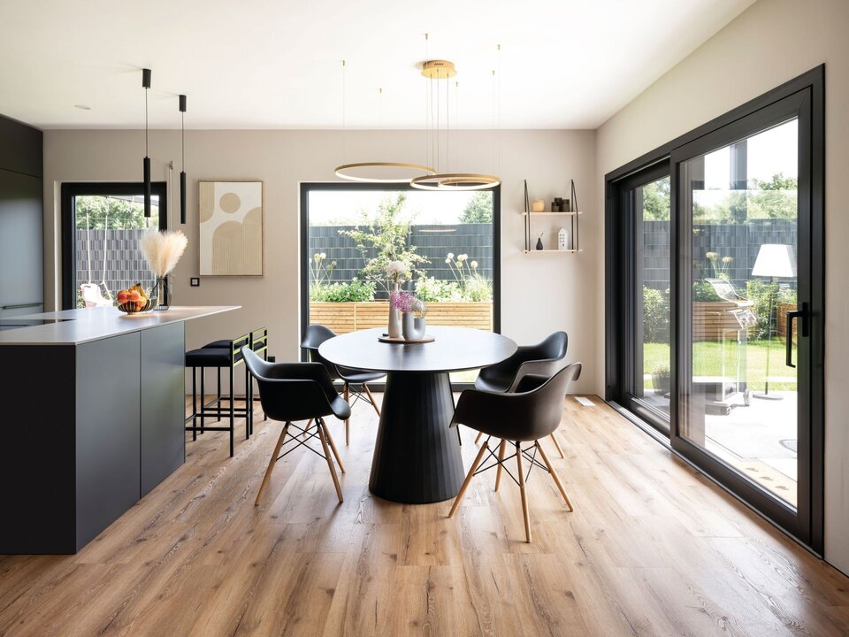 Heller Essbereich mit rundem schwarzen Tisch, vier schwarzen Stühlen und großem Fenster zur Terrasse. Offene Küche mit dunklen Fronten und heller Arbeitsplatte, Pendelleuchten über dem Tisch und Holzfußboden in Eichenoptik.