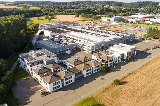 Luftaufnahme des Firmensitzes von FingerHaus in Frankenberg (Eder) mit Verwaltungsgebäuden und Produktionshalle, auf deren Dach zahlreiche Solarmodule installiert sind, umgeben von Feldern und Grünflächen.