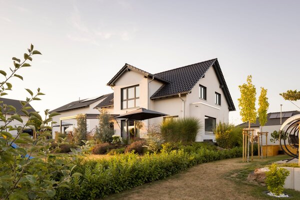 Außenansicht eines Einfamilienhauses SENTO 403 mit Satteldach, Gartenanlage, Sonnenschirm und bepflanzten Beeten im Abendlicht