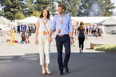 Ein Paar geht über das Veranstaltungsgelände des FingerHaus-Bauinfotags bei sonnigem Wetter. Beide tragen Besucherausweise um den Hals, im Hintergrund sind weitere Gäste und Informationsstände zu sehen.