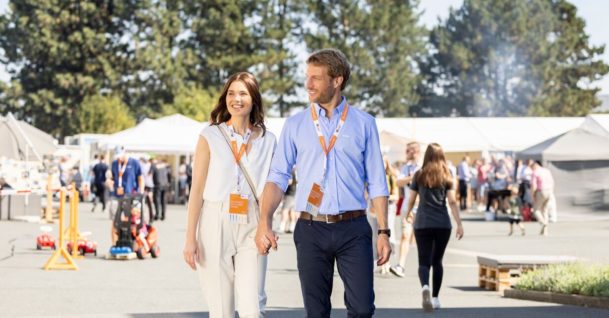 Ein Paar geht über das Veranstaltungsgelände des FingerHaus-Bauinfotags bei sonnigem Wetter. Beide tragen Besucherausweise um den Hals, im Hintergrund sind weitere Gäste und Informationsstände zu sehen.