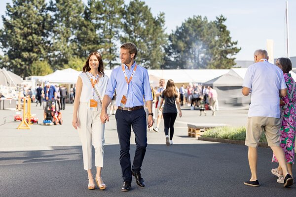 Ein Paar geht über das Veranstaltungsgelände des FingerHaus-Bauinfotags bei sonnigem Wetter. Beide tragen Besucherausweise um den Hals, im Hintergrund sind weitere Gäste und Informationsstände zu sehen.