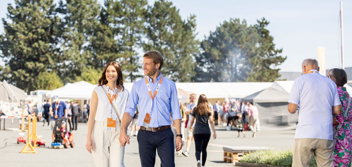 Ein Paar geht über das Veranstaltungsgelände des FingerHaus-Bauinfotags bei sonnigem Wetter. Beide tragen Besucherausweise um den Hals, im Hintergrund sind weitere Gäste und Informationsstände zu sehen.