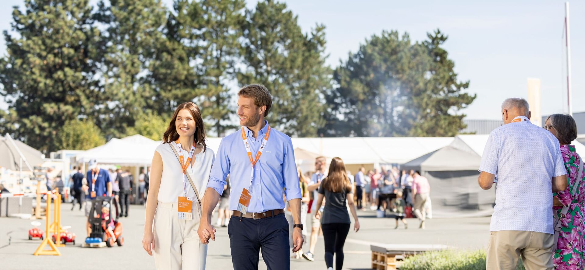 Ein Paar geht über das Veranstaltungsgelände des FingerHaus-Bauinfotags bei sonnigem Wetter. Beide tragen Besucherausweise um den Hals, im Hintergrund sind weitere Gäste und Informationsstände zu sehen.