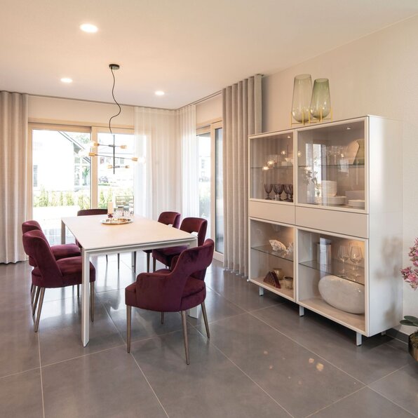 Essbereich im FingerHaus-Musterhaus MEDLEY 300 B in Frankenberg mit weißem Esstisch, sechs gepolsterten Stühlen in Bordeaux, großformatigen grauen Bodenfliesen, Glasvitrine mit Geschirr, bodentiefen Fenstern mit Vorhängen und moderner Pendelleuchte.