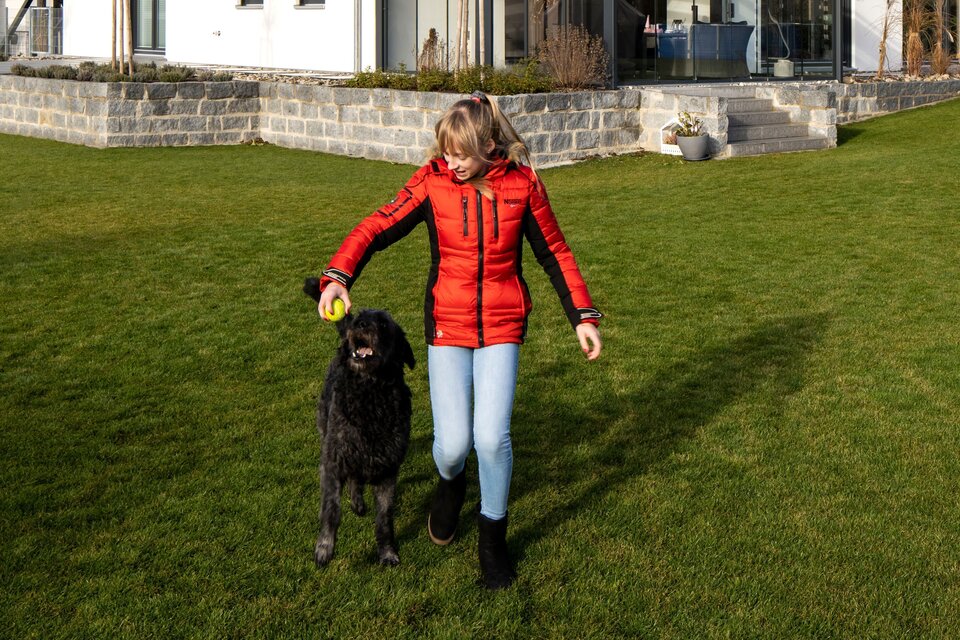Mädchen spielt mit einem schwarzen Hund auf der großen Rasenfläche vor einem modernen zweigeschossigen FingerHaus
