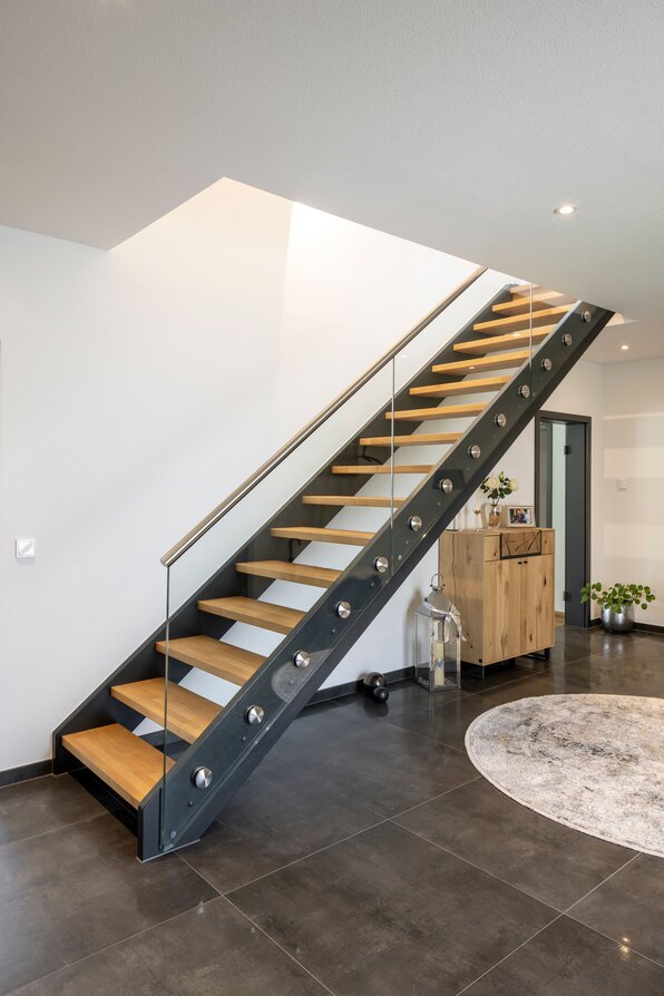 Moderne Treppe in einem frei geplanten FingerHaus mit offenen Holzstufen, seitlicher Stahlkonstruktion und Glasgeländer. Der Eingangsbereich ist hell gestaltet und mit eleganten Fliesen ausgelegt.