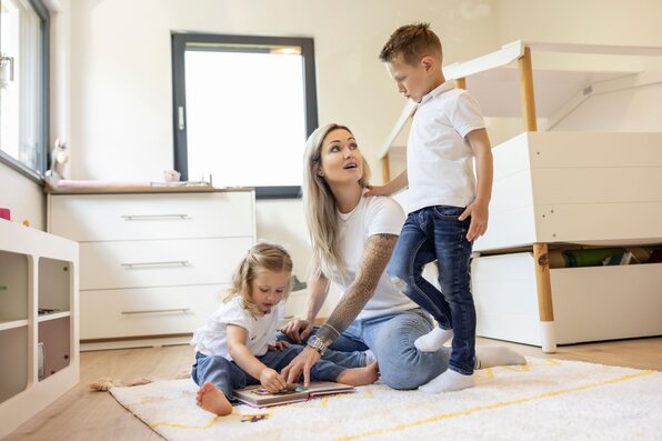 Eine Mutter sitzt mit ihren zwei Kindern im hellen Kinderzimmer eines modernen FingerHaus-Hauses, sie lesen gemeinsam auf dem Teppich und genießen die Zeit zusammen.