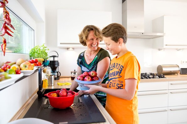 Eine Frau und ein Junge waschen gemeinsam frisches Obst in einer modernen, hellen Küche mit Holzarbeitsplatte.