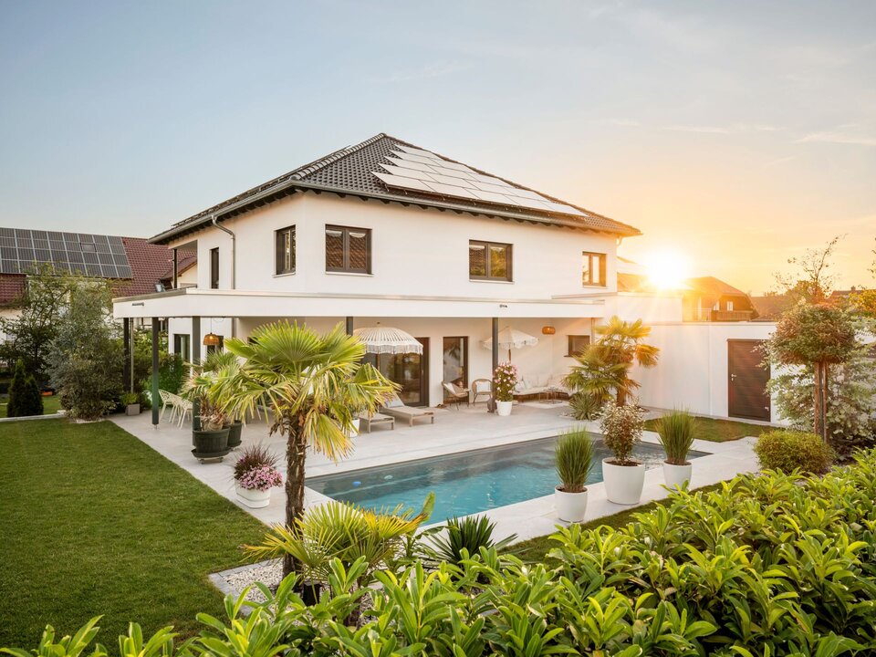 Frei geplante Stadtvilla mit überdachter Terrasse, Swimmingpool, Photovoltaikanlage und mediterran gestalteter Gartenanlage bei Sonnenuntergang.