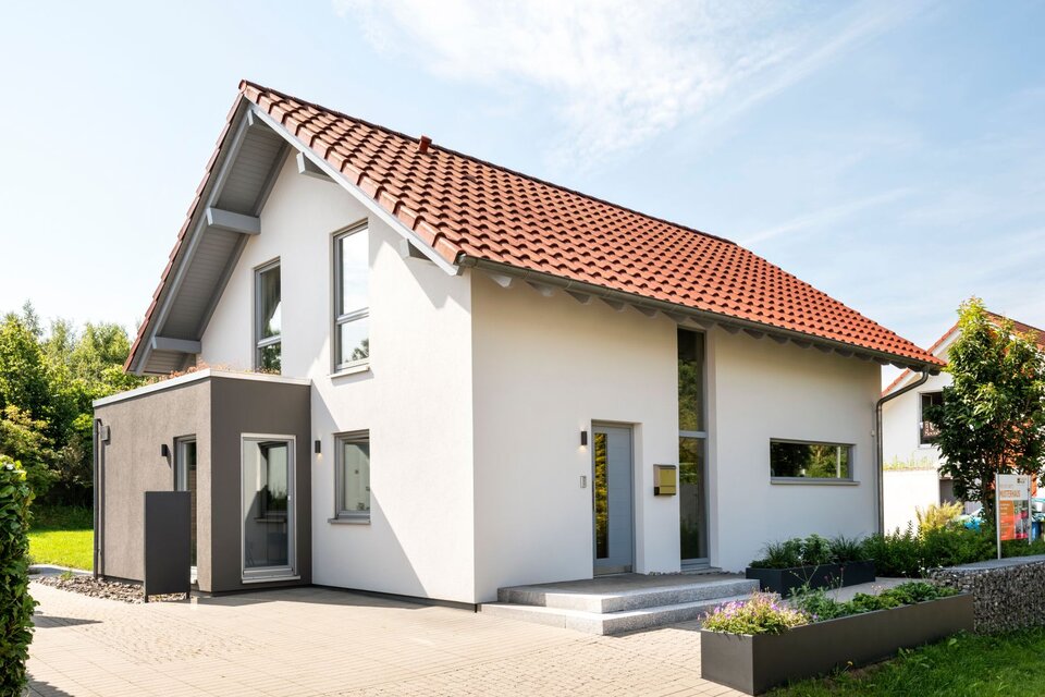 Außenansicht eines frei geplanten Finger-Haus Musterhauses in Marburg mit weißer Putzfassade, rotem Satteldach und dunkelgrauem Anbau; davor gepflasterte Terrasse, Hochbeete mit Bepflanzung, Rasenfläche und eine FingerHaus-Fahne am Grundstück.