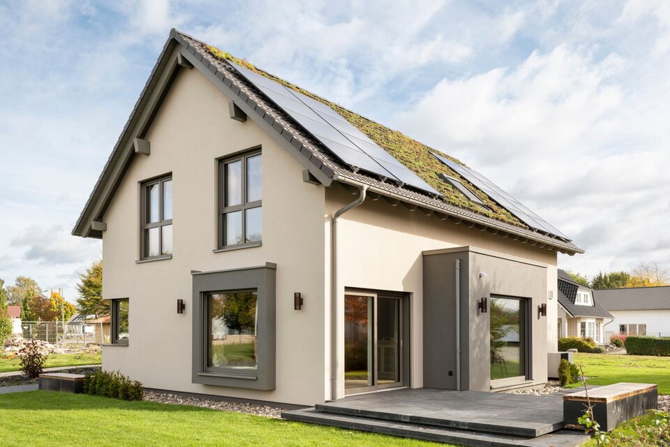 Rückansicht des Musterhauses JUNO 300 in Mülheim-Kärlich mit begrüntem Dach, Solarpaneelen und Terrasse.
