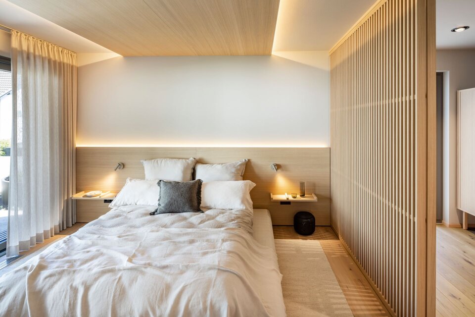 Schlafzimmer im Musterhaus NIVO »Sally Edition« im Musterhauspark Frankenberg mit Doppelbett, indirekter Beleuchtung, Holzlamellenwand und offenem Übergang zur Ankleide.