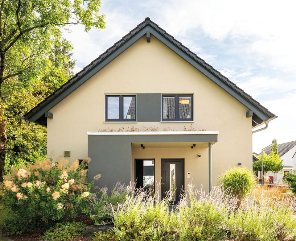 Eingangsbereich des Musterhauses NEO in Wuppertal mit überdachter Haustür, heller Fassade und umrahmt von moderner Gartenbepflanzung.