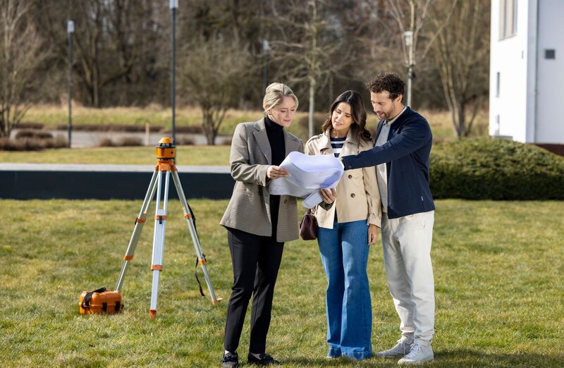 Eine Architektin zeigt einem Paar auf einem Grundstück Baupläne; im Hintergrund steht ein Vermessungsgerät auf einem Stativ.
