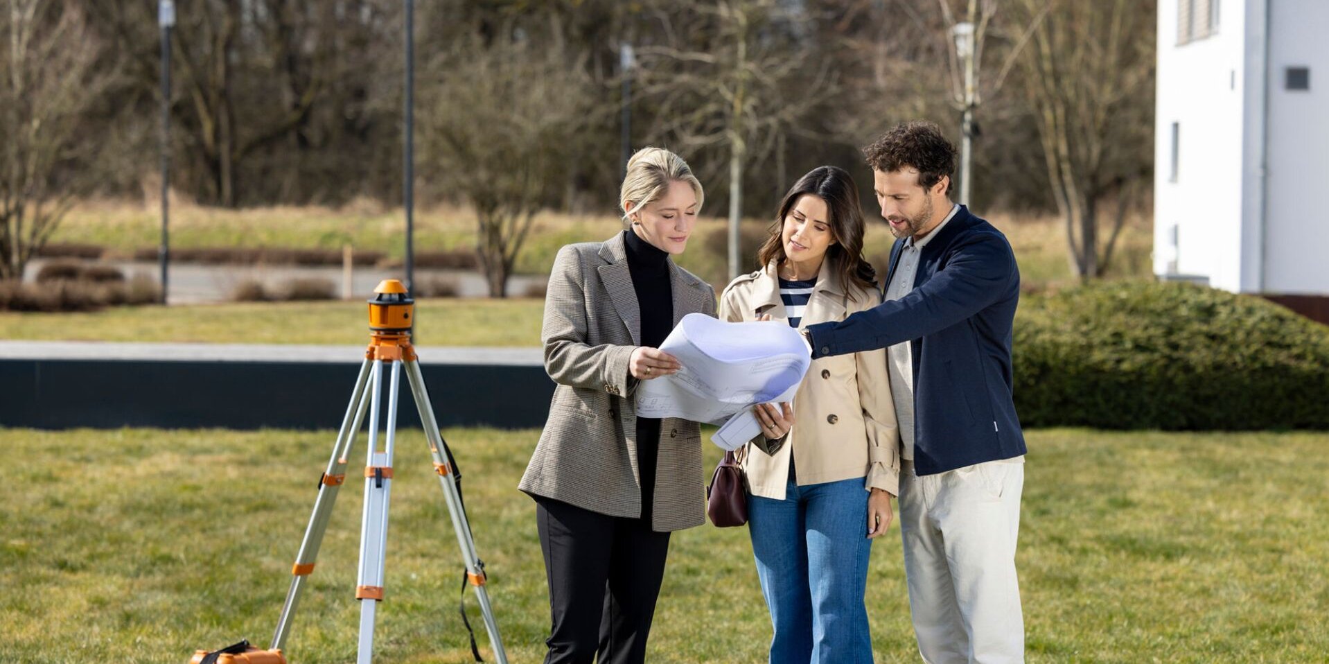 Eine Architektin zeigt einem Paar auf einem Grundstück Baupläne; im Hintergrund steht ein Vermessungsgerät auf einem Stativ.
