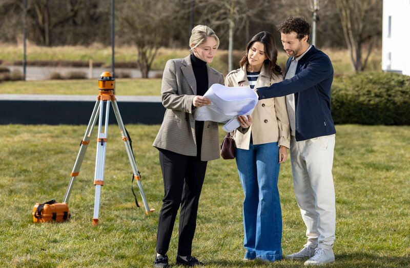 Eine Architektin zeigt einem Paar auf einem Grundstück Baupläne; im Hintergrund steht ein Vermessungsgerät auf einem Stativ.