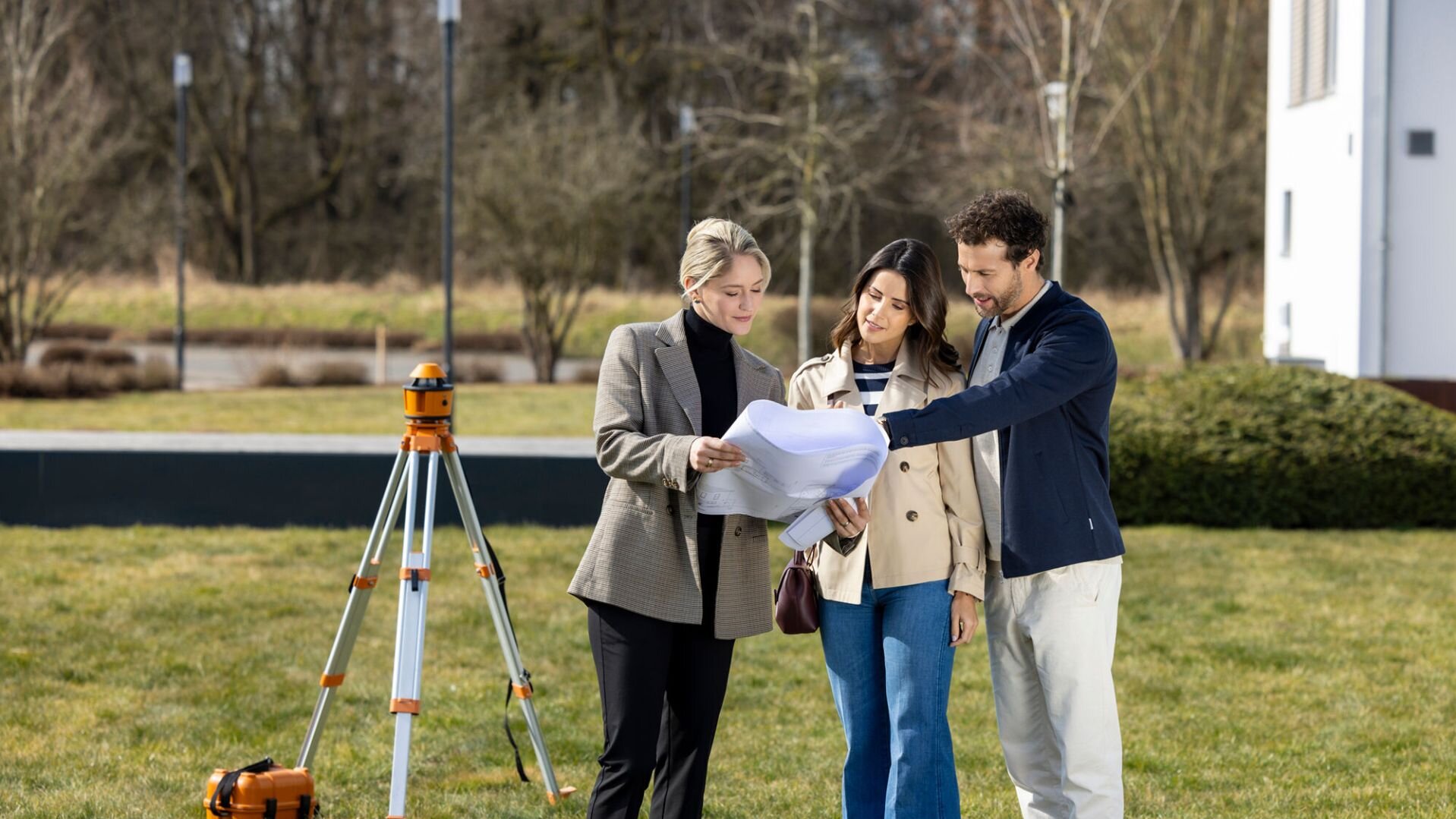 Eine Architektin zeigt einem Paar auf einem Grundstück Baupläne; im Hintergrund steht ein Vermessungsgerät auf einem Stativ.
