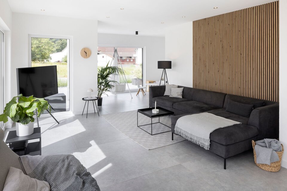 Modernes Wohnzimmer in einem frei geplanten FingerHaus. Großzügiger, heller Raum mit grauem Ecksofa, Holzpaneel-Wand, großen Fenstern und Blick in den Garten.