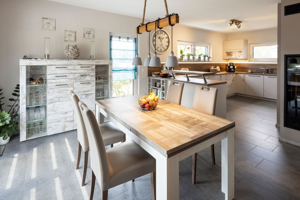 Essbereich mit hellem Holztisch und sechs Stühlen, angrenzend an eine offene Küche. Holzoptik, grauer Fliesenboden und große Fenster sorgen für ein freundliches, modernes Ambiente.