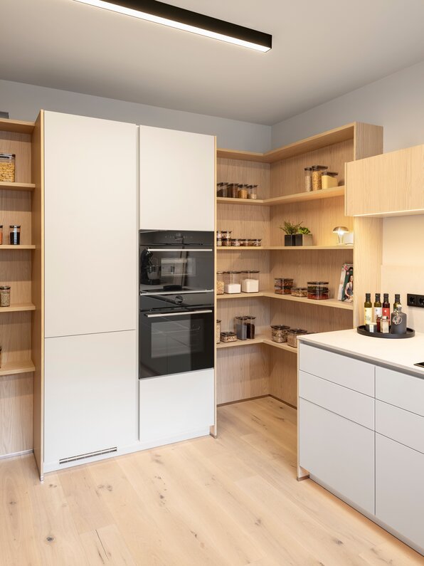 Küche im FingerHaus Musterhaus NIVO in Frankenberg mit Hochschrank, zwei Einbauöfen und offenen Holzregalen mit Vorratsgläsern.