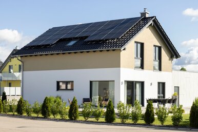 Einfamilienhaus des Hausmodells NEO 211 von FingerHaus mit Satteldach und Photovoltaikanlage bei Tageslicht, helle Putzfassade mit zweifarbiger Gestaltung, bodentiefe Fenster, Terrasse mit Sitzmöbeln und gepflegte Gartenbepflanzung.