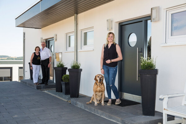 Drei Personen und ein Hund vor den separaten Hauseingängen eines frei geplanten Finger-Hauses mit Einliegerwohnung.