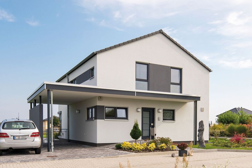 Außenansicht des Finger-Haus Musterhauses MEDLEY 300 B in Berlin/Werder: zweigeschossiges Haus mit weißer Fassade und grauen Akzentflächen, Satteldach und Carport; davor gepflasterte Einfahrt mit geparktem Auto, Beetbepflanzung, Rasen und Bäume unter blauem Himmel.