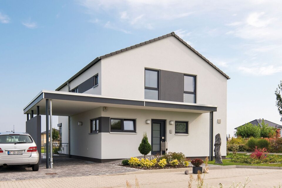 Außenansicht des Finger-Haus Musterhauses MEDLEY 300 B in Berlin/Werder: zweigeschossiges Haus mit weißer Fassade und grauen Akzentflächen, Satteldach und Carport; davor gepflasterte Einfahrt mit geparktem Auto, Beetbepflanzung, Rasen und Bäume unter blauem Himmel.