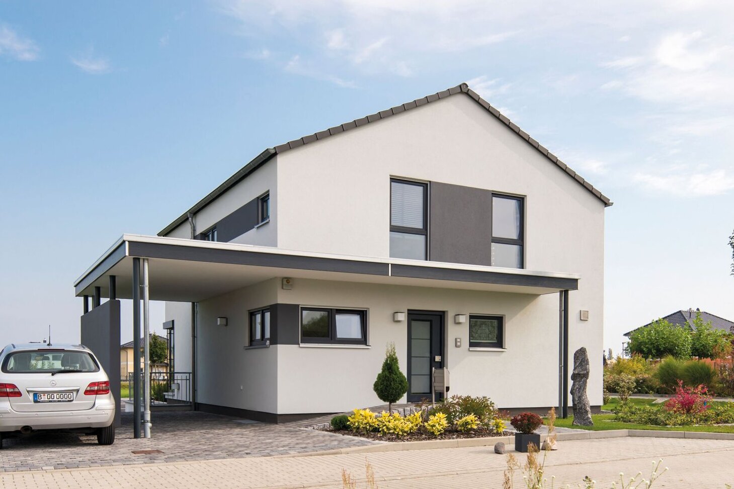 Außenansicht des Finger-Haus Musterhauses MEDLEY 300 B in Berlin/Werder: zweigeschossiges Haus mit weißer Fassade und grauen Akzentflächen, Satteldach und Carport; davor gepflasterte Einfahrt mit geparktem Auto, Beetbepflanzung, Rasen und Bäume unter blauem Himmel.